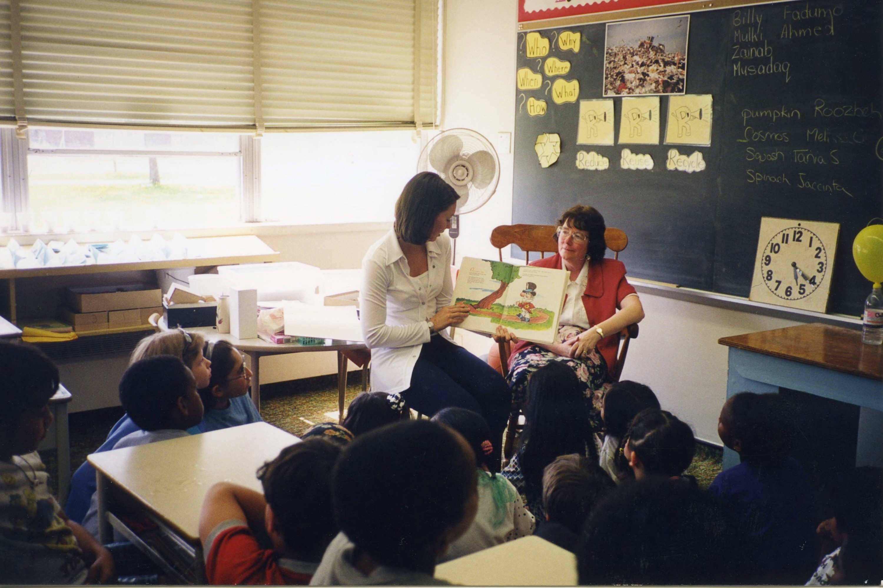Professor Frize giving a presentation to students in primary school in Ottawa, she sits in a chair and is accompanied by a teacher who is holding a book up to the class