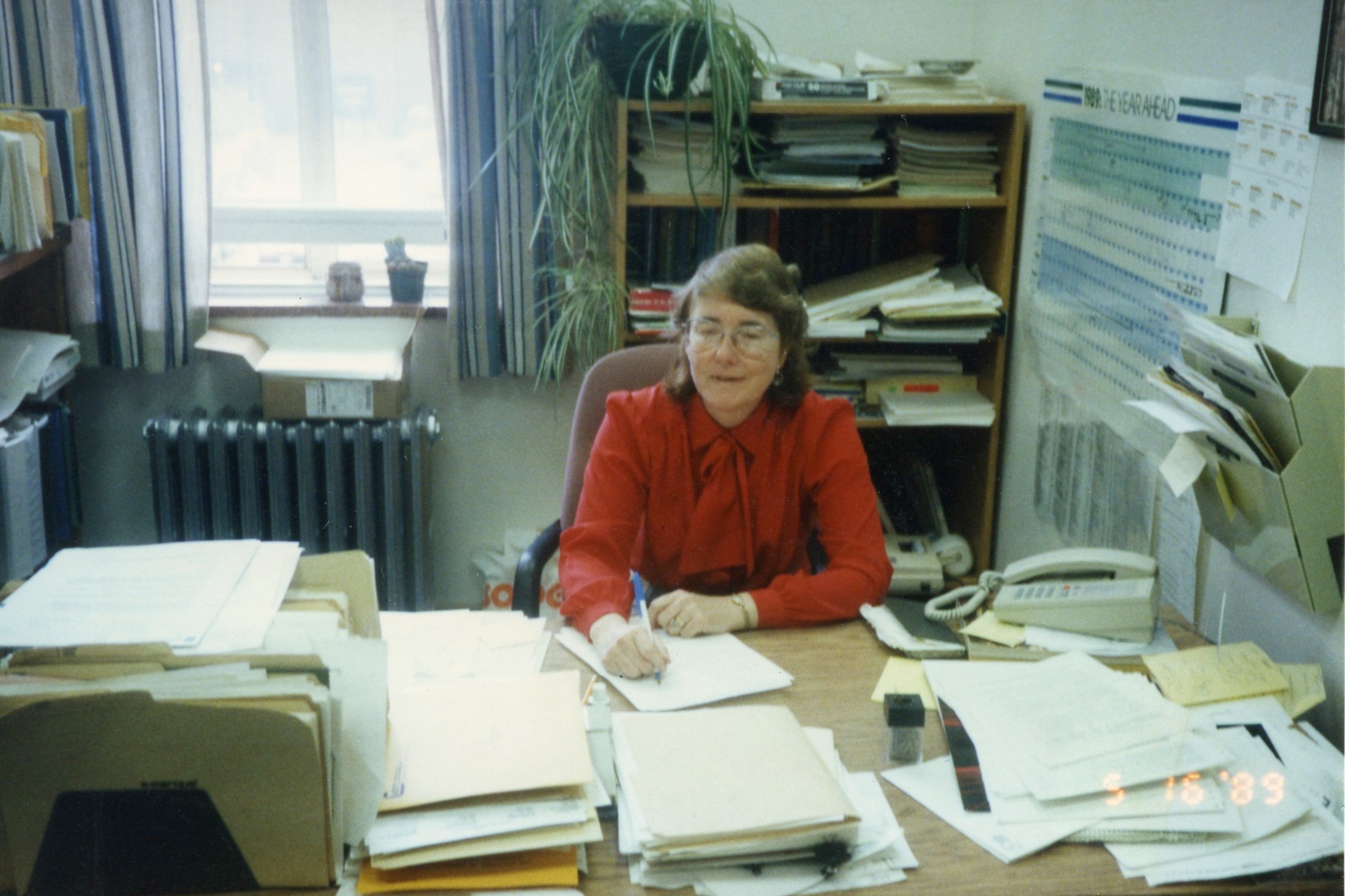 Professor Frize sitting at a desk in a small room, the desk is covered in stacks of paper and there are a set of shelves and a window behind her. The image is dated as 1989.