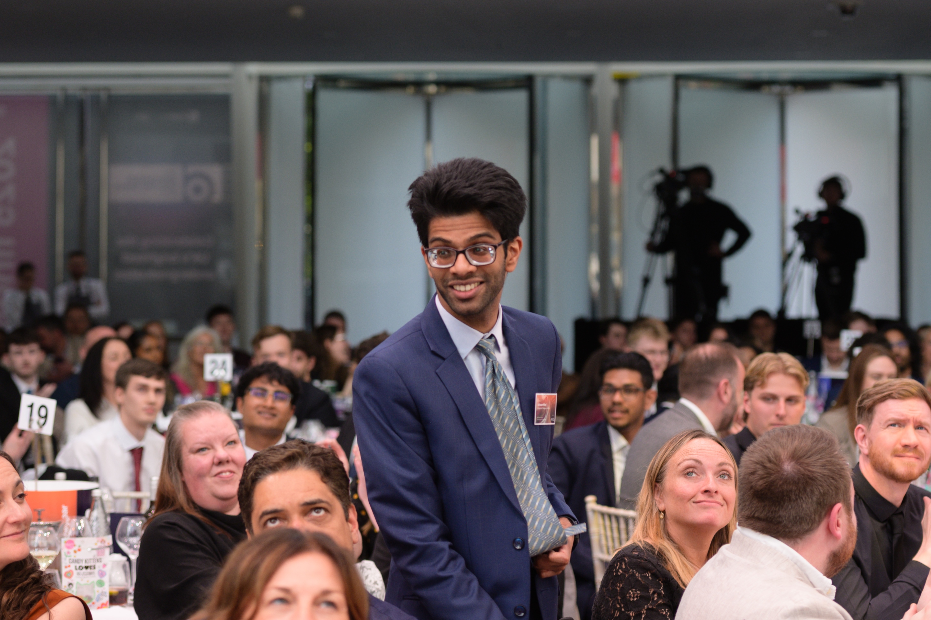 Photo of Eshaan Niraj standing to accept his award, there are other people sat around him, most looking towards the camera